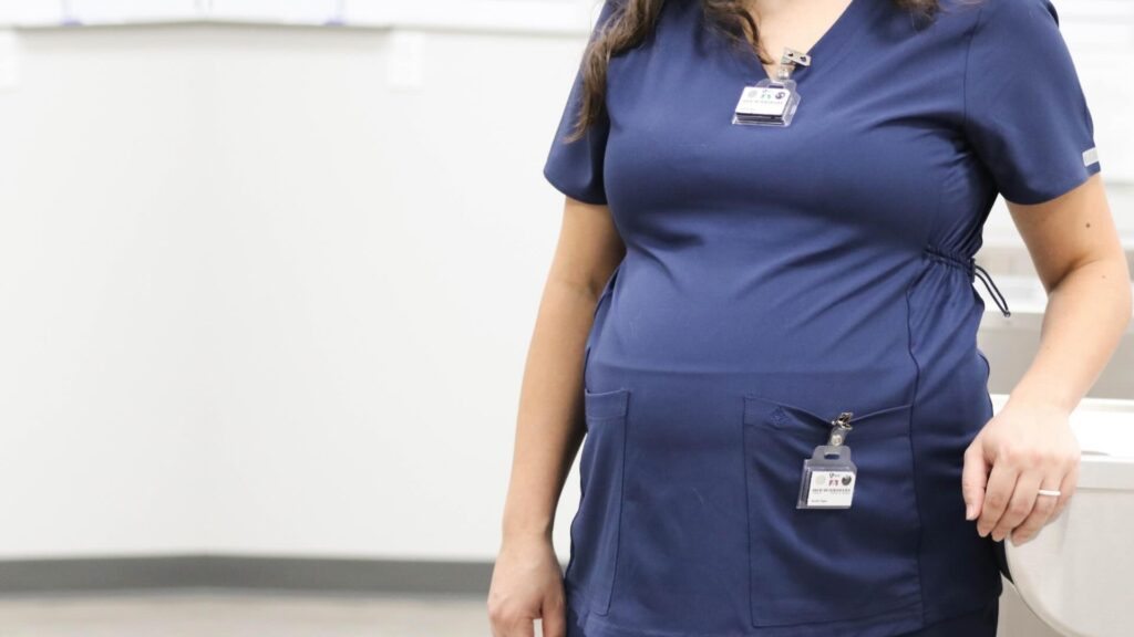 A pregnant woman wears a TLD dosimeter at the collar of her dark blue scrubs with another TLD dosimeter at her waist for fetal monitoring.