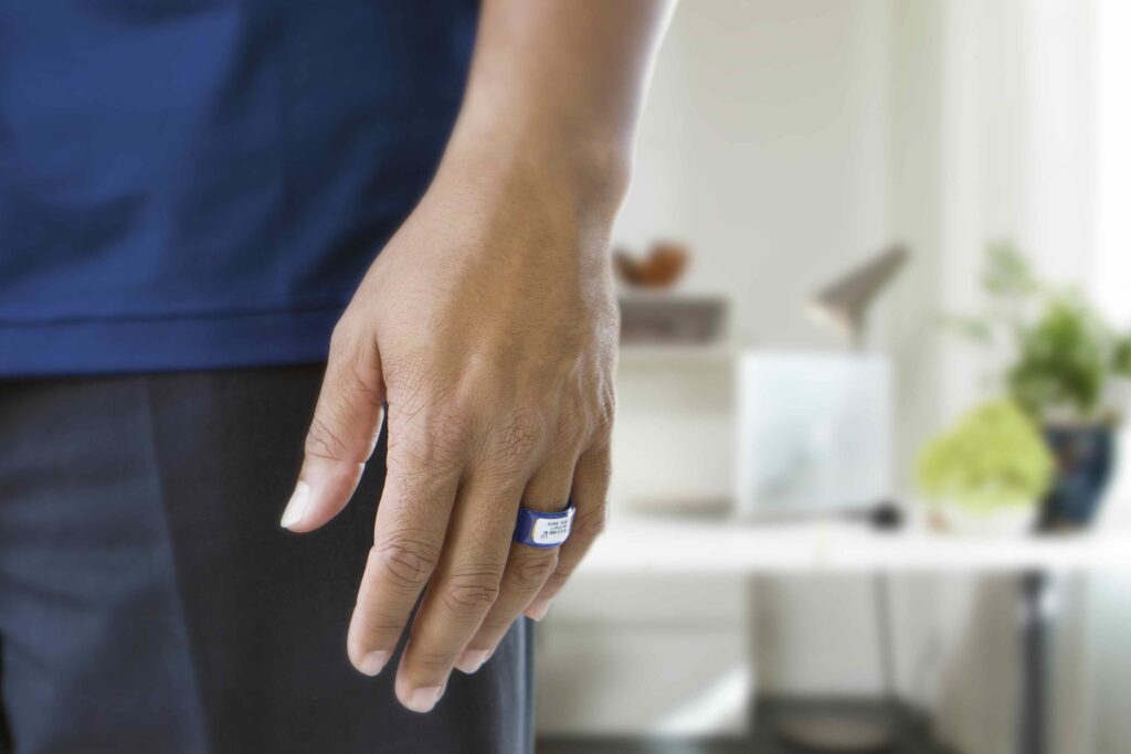 Close-up of a person’s hand wearing a sleek and modern TLD ring. The background reveals a blurred indoor setting with a desk, lamp, and plants, drawing focus to the innovative ring.