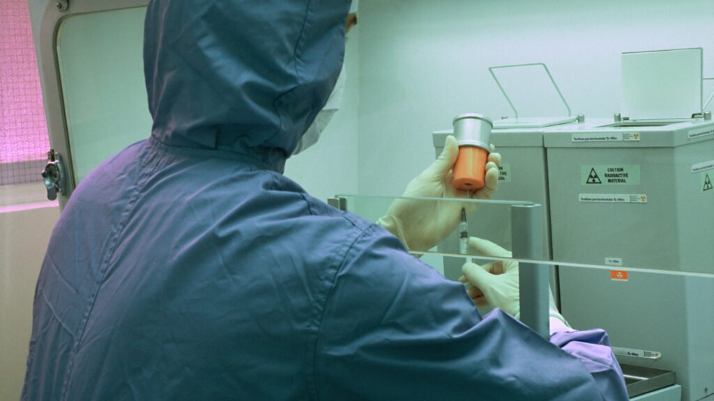 An employee carefully handles radioactive materials behind a barrier while wearing PPE.