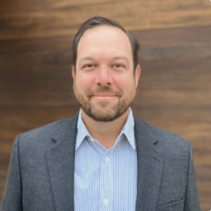 Matt McColgan, a man with a beard, wearing a blue striped shirt and a dark blazer, stands in front of a blurred wooden background, smiling at the camera.