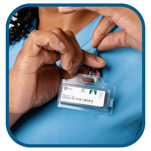 A person in a blue uniform adjusting a name badge clipped to their shirt. The badge has a barcode, logo, and an icon of a heart and stethoscope, suggesting a medical or healthcare setting. The persons face is not visible.