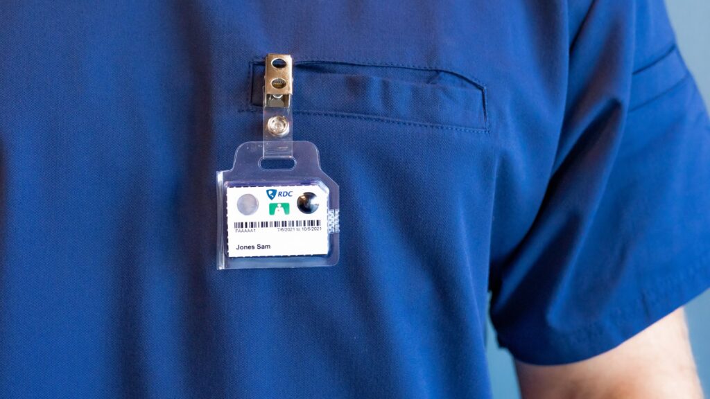 An employee wears a TLD badge on his scrubs for dosimetry monitoring during an imaging session for a patient.