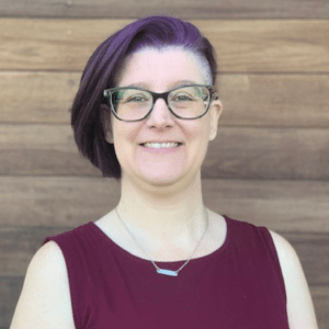 A person with short purple hair and glasses, smiling in a sleeveless burgundy top, stands confidently against a wooden wall background, embodying the vibrant spirit of UT Health.
