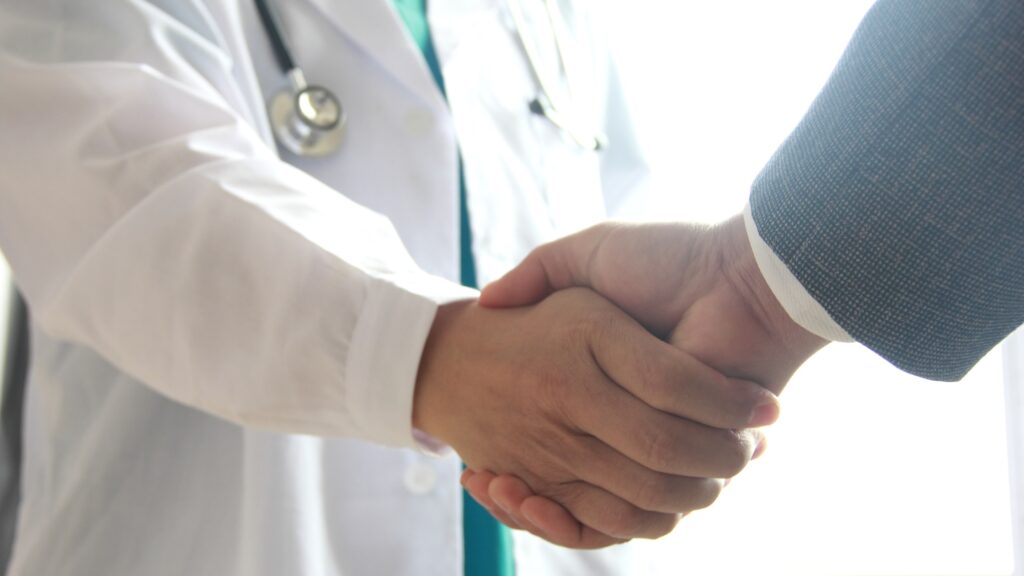 A doctor in a white coat and a business professional shake hands.