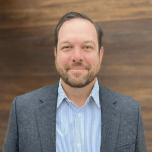 A man with short hair and a beard, wearing a blue striped shirt and a gray blazer, is smiling slightly while standing in front of a wooden wall at UT Health.