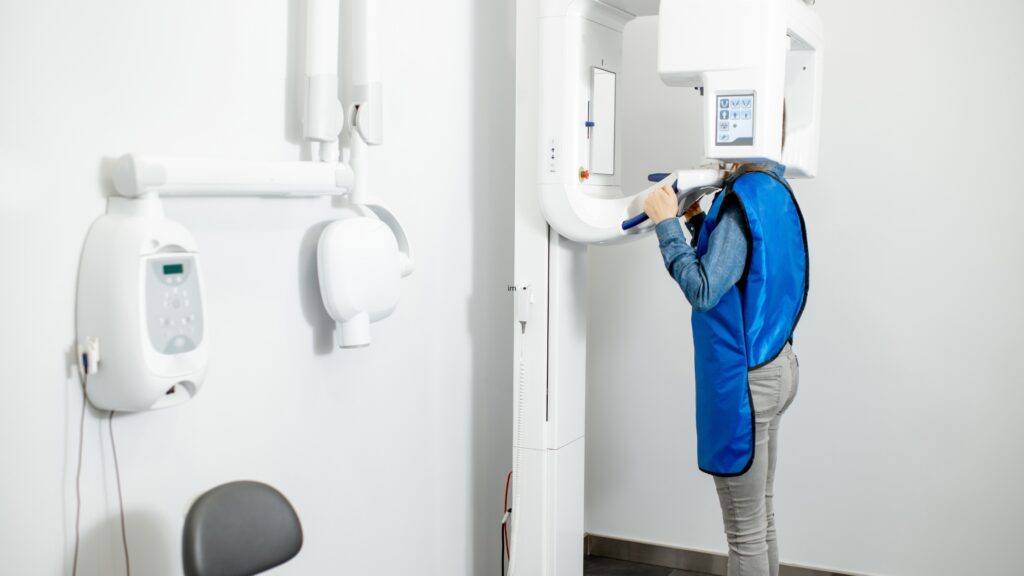 A patient receives a dental X-ray while staff distance themselves in the other room.