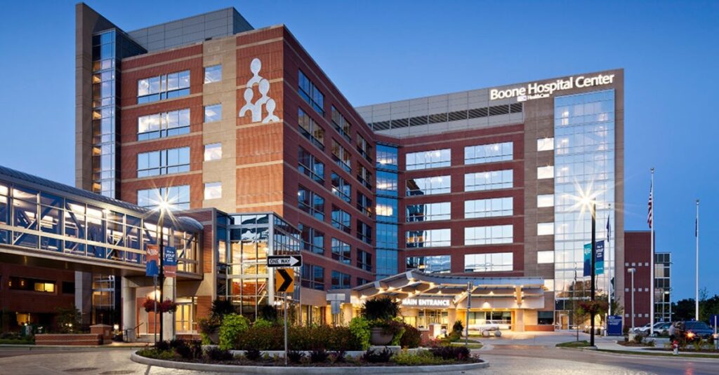 Night view of a modern hospital building with large glass windows and a sign reading Boone Hospital Center. The main entrance is well-lit, and the surrounding area is landscaped with greenery and pathways.