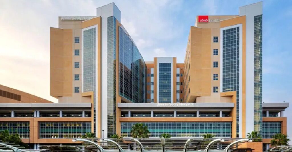 A modern hospital building with multiple stories, featuring a blend of glass and brown facade. The architecture includes tall vertical sections and a large, central glass atrium. The name Jennie Sealy Hospital is displayed prominently.