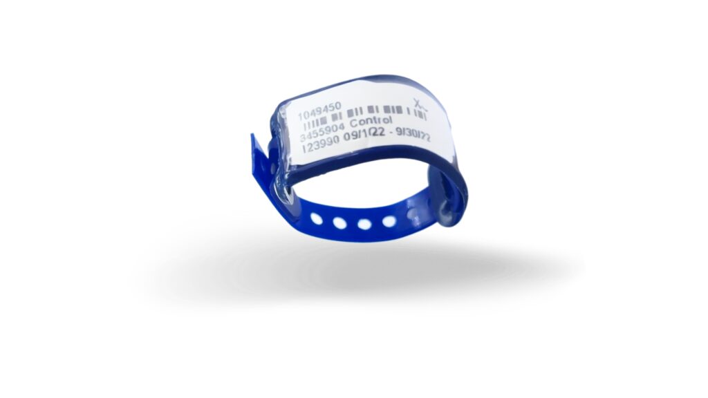 A blue TLD ring with a white label displays barcodes and alphanumeric information, partially legible for extremity monitoring. The band is curled, showcasing circular holes for adjustment against the clean white background.