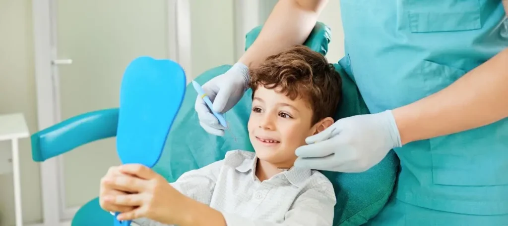 A young boy sits in a dental chair, smiling as he holds a blue mirror. Beside him, a dentist in gloves and scrubs is poised with dental tools, ensuring everything is perfect. In this bright, modern dental office, cutting-edge techniques like dosimetry monitoring enhance the care provided.