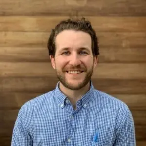 Michael Boches with short, curly hair and a beard is smiling while wearing a blue and white checkered shirt. He is standing in front of a wooden background.