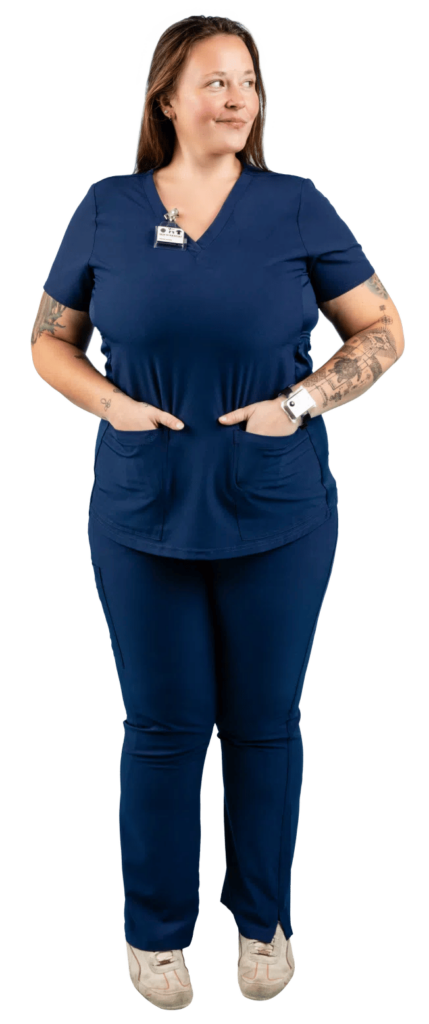 A person wearing a navy blue scrub suit with a name badge and a watch stands with hands in pockets. They have long hair and visible tattoos on their arms. They are smiling and looking to the side.