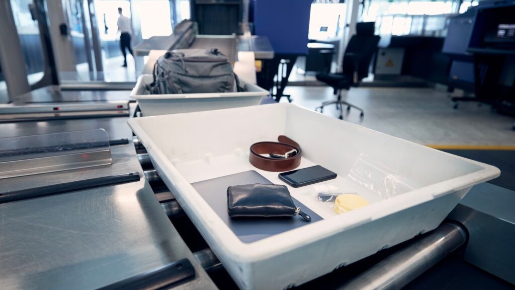 A white security bin at an airport checkpoint contains a wallet, smartwatch, smartphone, belt, and small clear bag. A backpack is visible in another bin ahead on the conveyor belt near metal detectors.