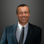 Alastair Watson, dressed in a dark suit and striped tie, smiles at the camera against a plain, dark gray background.
