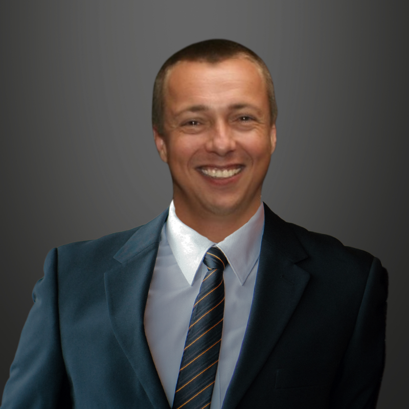 Alastair Watson, dressed in a dark suit and striped tie, smiles at the camera against a plain, dark gray background.