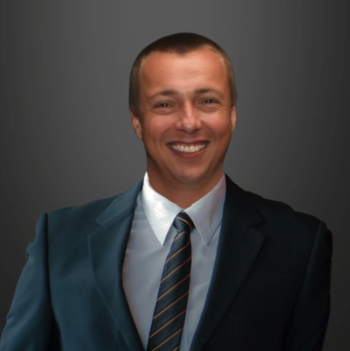 Alastair Watson, dressed in a dark suit and striped tie, smiles at the camera against a plain, dark gray background.