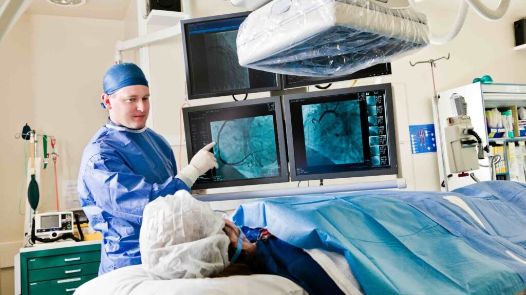 A medical professional in surgical scrubs points at medical scans on monitors, reviewing the patient’s radiation dose while attending to a patient lying on an operating table, covered with a blue sheet in a modern hospital room with medical imaging equipment.