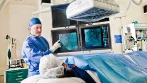 A medical professional in surgical scrubs points at medical scans on monitors, reviewing the patient’s radiation dose while attending to a patient lying on an operating table, covered with a blue sheet in a modern hospital room with medical imaging equipment.
