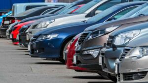 A row of parked cars in various colors and models, lined up closely side by side in an outdoor parking lot, primed for dosimeter fade in the summer heat.
