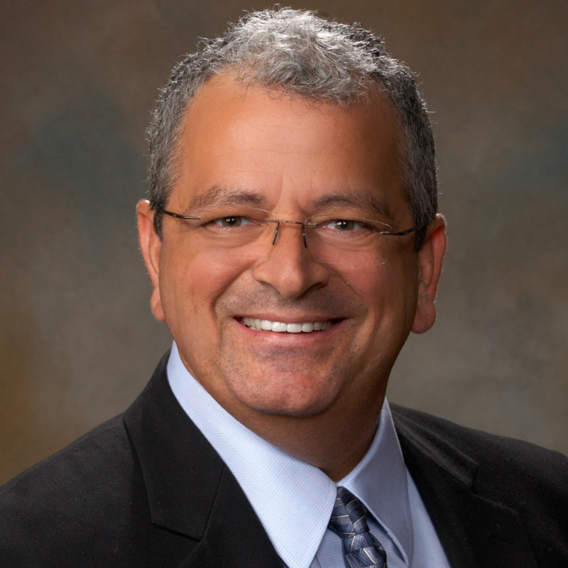 Chuck Nagel, a smiling man with short gray hair, wearing glasses, a dark suit jacket, light blue shirt, and patterned tie, poses in front of a blurred brown background.
