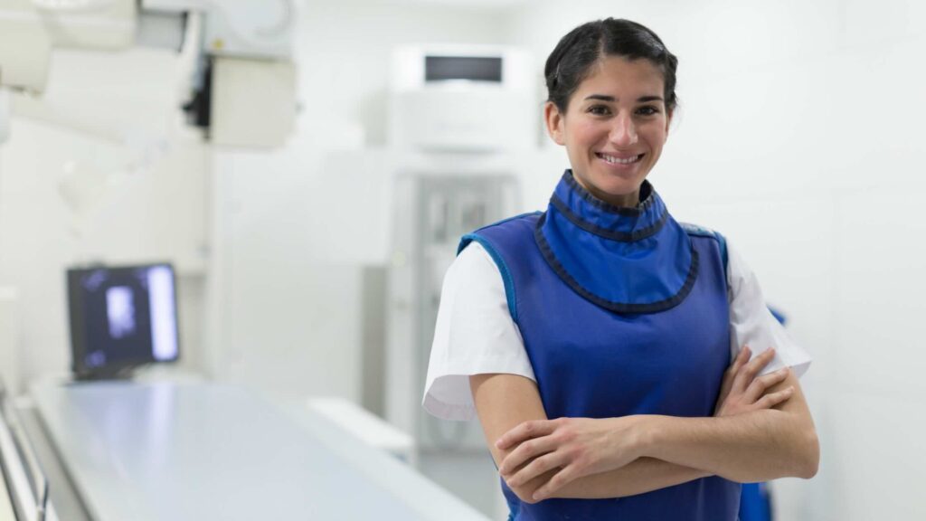 A smiling medical professional wearing a blue lead apron stands with arms crossed in a bright, modern radiology or X-ray room.