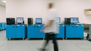 A person walks past a row of blue computer workstations, where depletion handling systems run smoothly. The clean, well-lit room features monitors and black towers; the person appears blurred from motion.