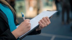 A person holding a clipboard and writing with a pen, wearing a blue shirt and dark jacket, with a blurred background.
