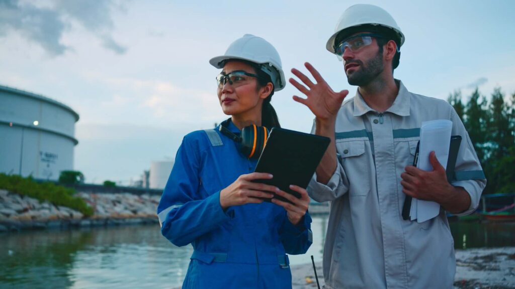 Two energy workers discuss plans at a nuclear site while wearing hardhats and carrying clipboards.