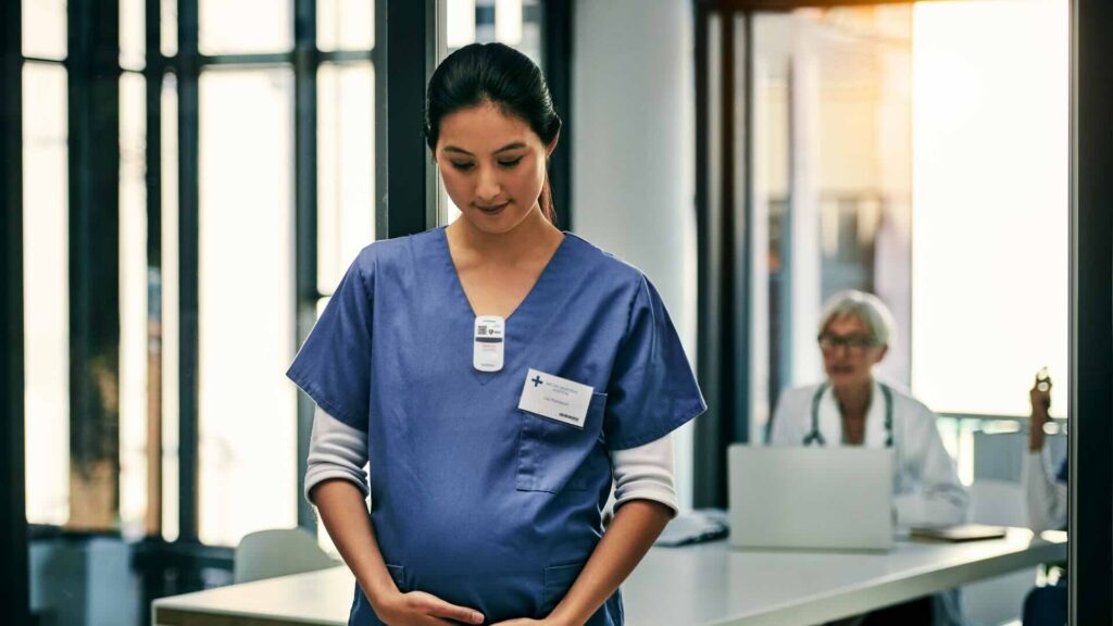 A tech in light blue scrubs holds her pregnant belly while wearing a digital dosimeter on the collar of her scrubs with a doctor sitting at a table working on a laptop in the background.