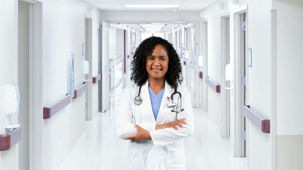 A doctor in light blue scrubs wears a white coat with an OSL dosimeter on the label and a stethoscope in the empty hallway of a hospital.