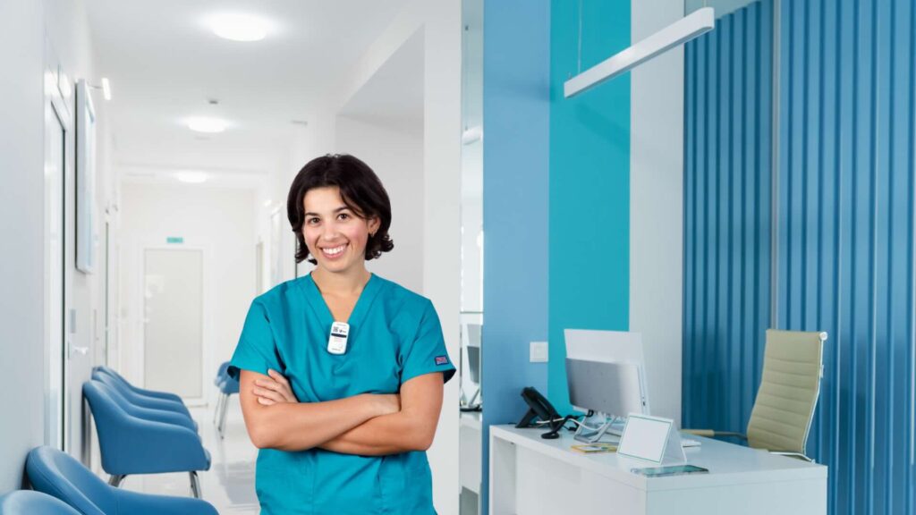 A medical professional in teal scrubs with short dark hair and her arms crossed across her chest wears a digital dosimeter on the collar of her scrubs with a waiting room in the background.