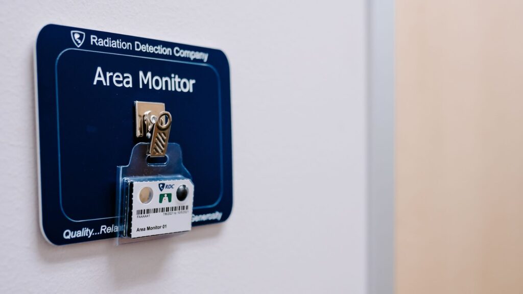 An area monitoring dosimeter badge is hanging on a badge clip mounted to a badge board against a dark blue background.