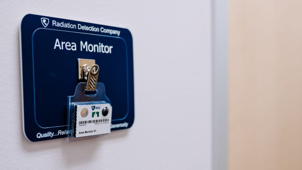 An area monitoring dosimeter badge is hanging on a badge clip mounted to a badge board against a dark blue background.