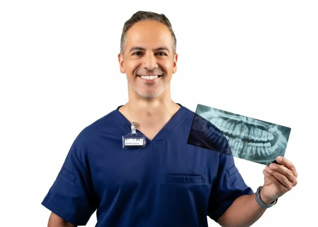 A smiling man in blue scrubs holding a dental X-ray, standing against a plain white background. He has a name badge clipped to his shirt.