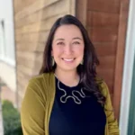 Amanda Smith, a woman with long brown hair wears a green cardigan and black shirt with gold necklace, smiling and standing in front of a wooden wall.