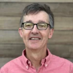 Headshot of Bruce Carmedelle wearing a soft red shirt and glasses outside of Radiation Detection Company with a wood background.