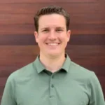 A person with short dark hair is smiling and wearing a green collared shirt, standing in front of a wooden wall background.