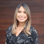 Michelle Uribe, a woman with long, straight hair and a center part smiles at the camera. She is wearing a black blouse with white leaf patterns. The background consists of wooden panels.