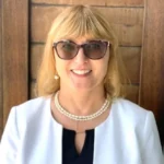 A woman with blonde hair and sunglasses grins effortlessly in front of a wooden backdrop. Her white blazer drapes over a black top, highlighted by a pearl necklace. Perhaps shes just closed a deal for a cutting-edge radiation detection company.