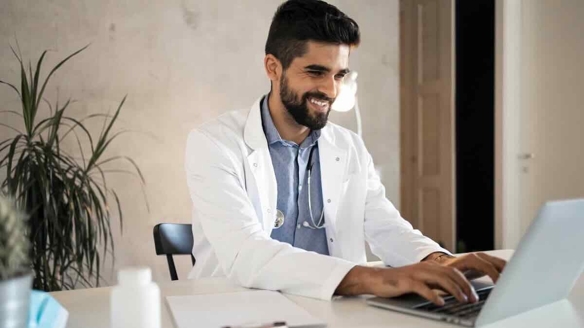 A smiling male doctor wearing a white coat experiences white-glove onboarding via his laptop, a stethoscope around his neck and medical supplies nearby.