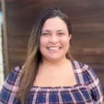 Vanessa Tardencilla, a woman with long hair smiles warmly at the camera. She is wearing a plaid shirt and standing in front of a wooden wall.