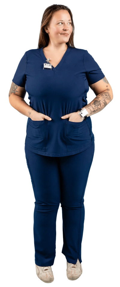 A person wearing a navy blue scrub suit with a name badge and a watch stands with hands in pockets. They have long hair and visible tattoos on their arms. They are smiling and looking to the side.