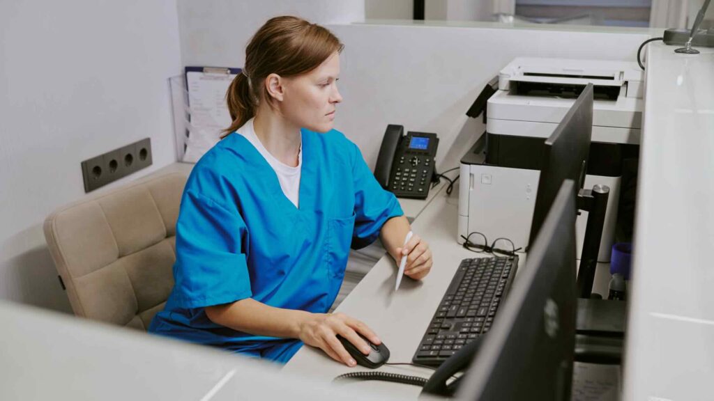 An imaging tech in blue scrubs researches dosimetry badge pricing at a desk.