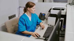An imaging tech in blue scrubs researches dosimetry badge pricing at a desk.